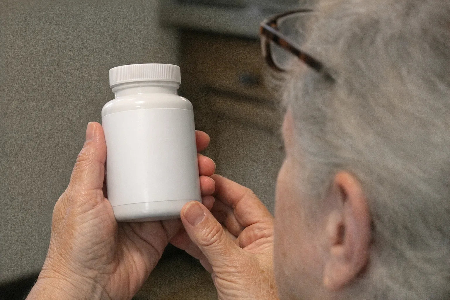 Older person's hands holding a plain supplement bottle at arm's length under kitchen light