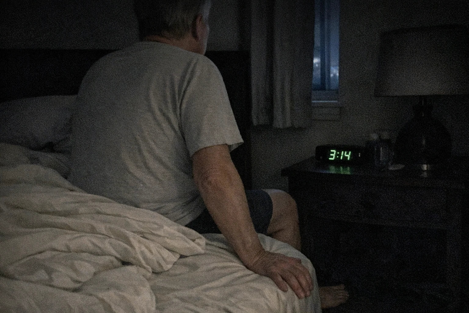 Phone photo of a dark bedroom at night, an older person sitting on the edge of the bed with a clock reading 3:14