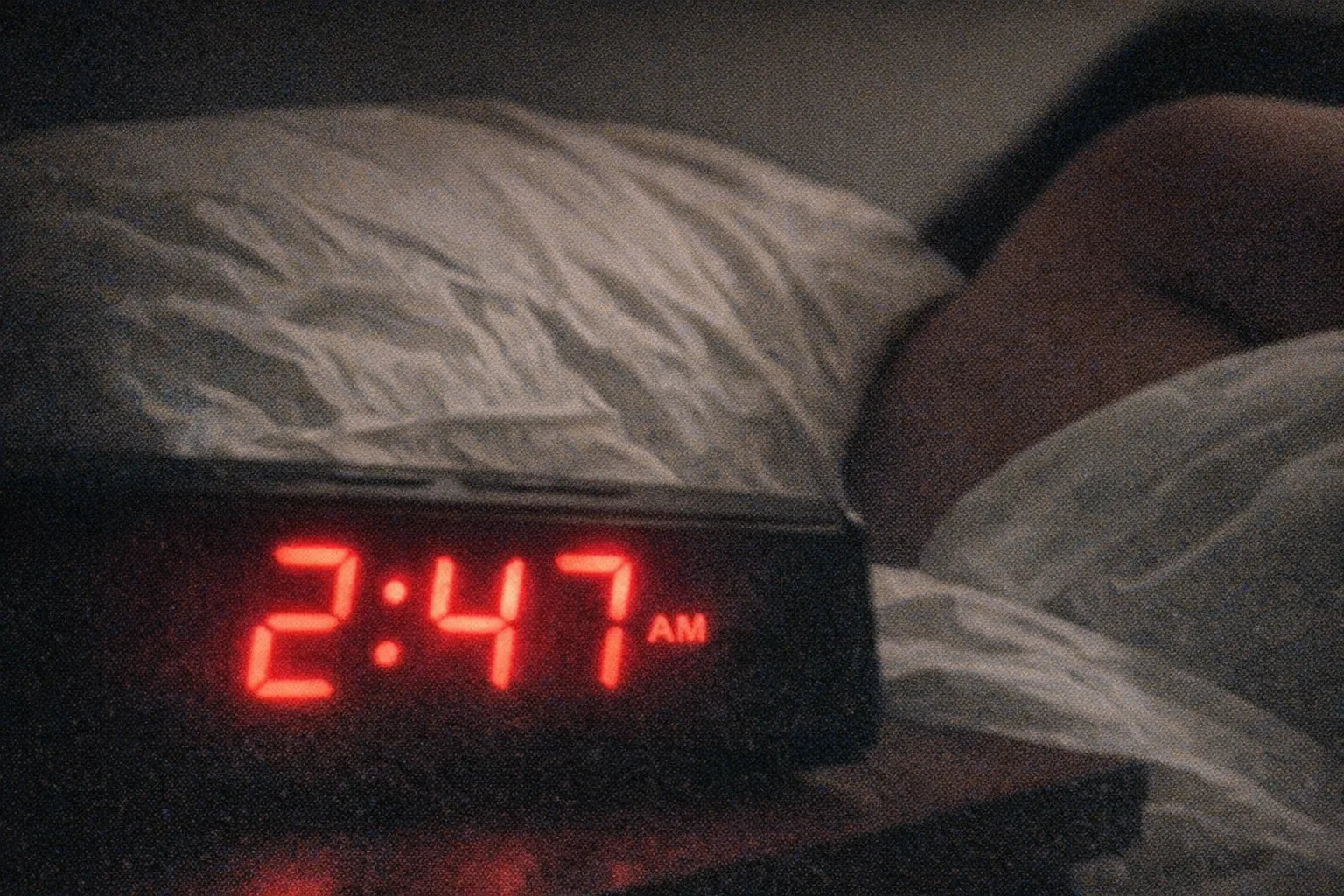 Close-up of a bedside clock reading 2:47 AM in red numerals with a rumpled pillow behind it