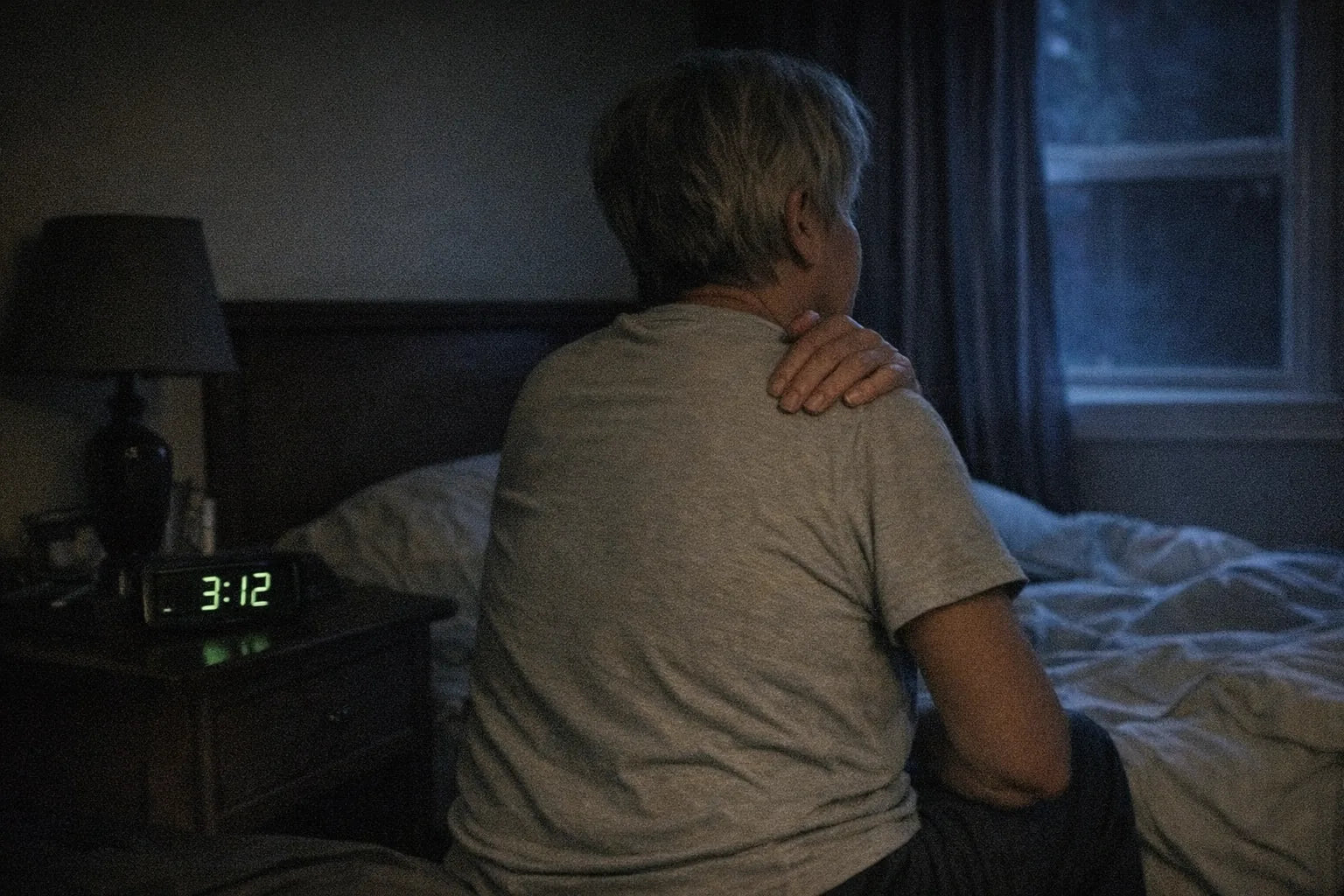 Phone photo of a dim bedroom at night, an older person sitting on the edge of the bed rubbing one shoulder, alarm clock reading 3:12