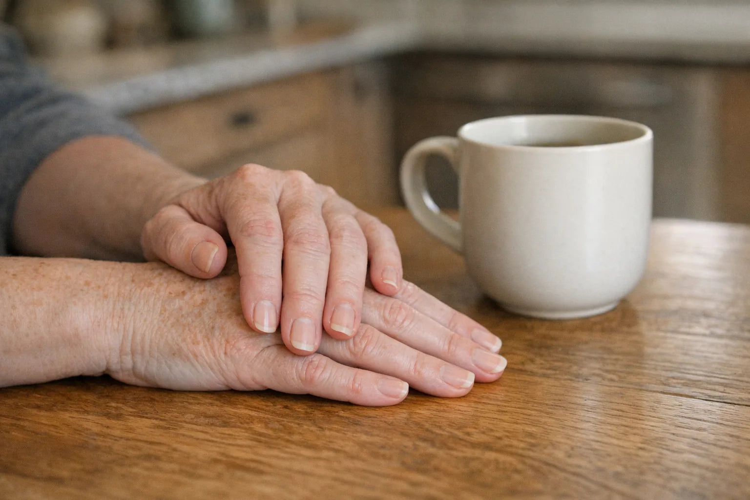 An older person's hands resting on a kitchen table in morning light with a mug beside them