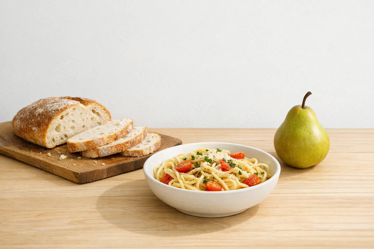 A bowl of pasta, a sliced loaf of bread and a pear on a wooden table